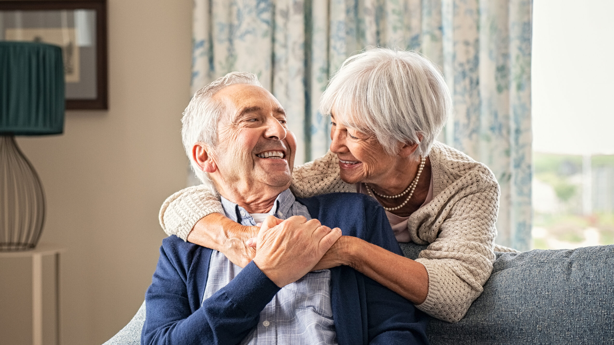 An older woman embraces an older man from behind. They are looking at each other and smiling.
